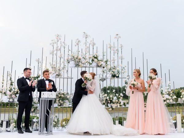 Front view of wedding ceremony outdoors, man in suits kissing woman in white puffy dress, while standing with bridesmaids and groomsmen near floral arch.